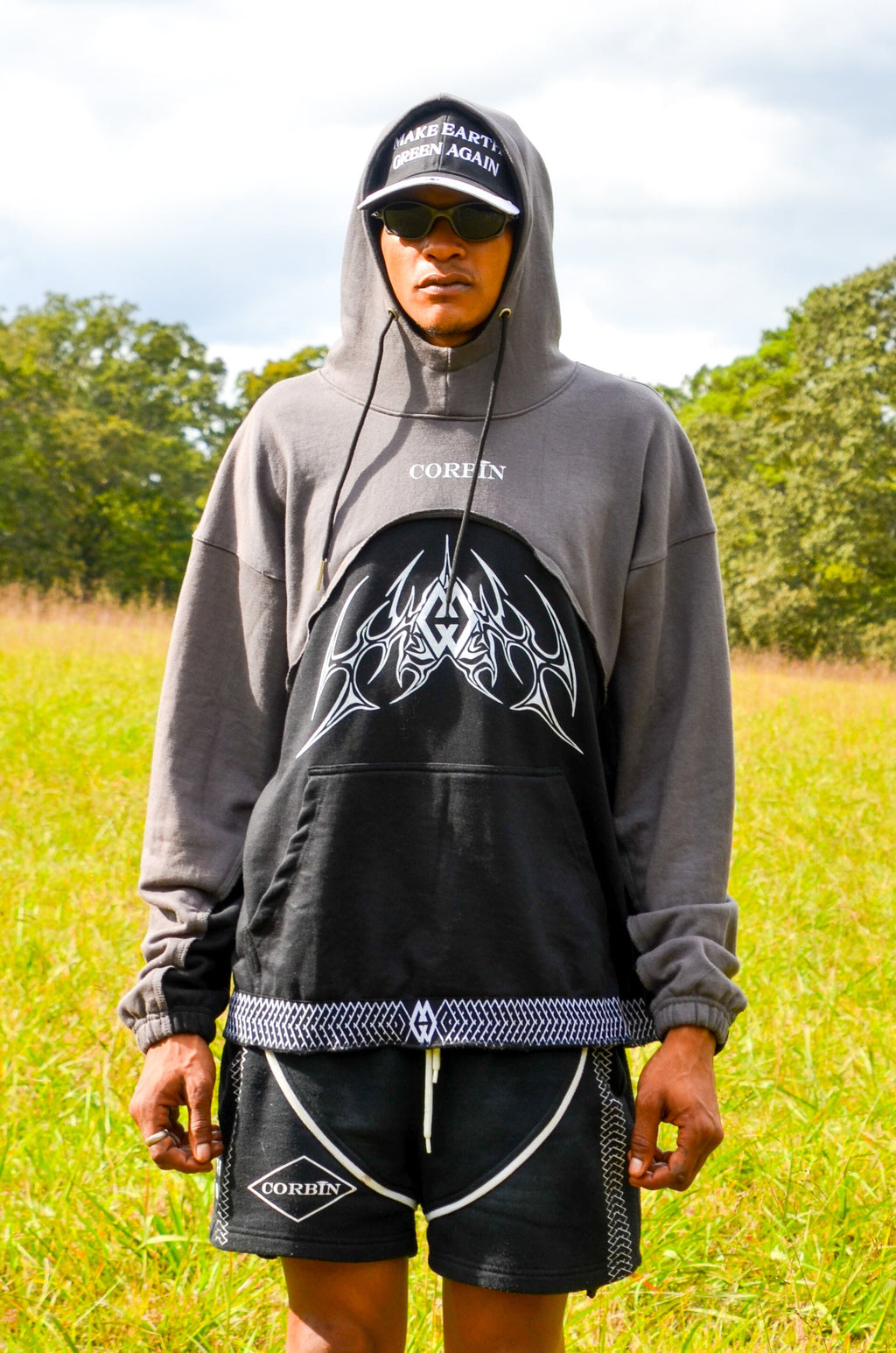 Person wearing a hoodie and shorts standing in a grassy field with trees in the background